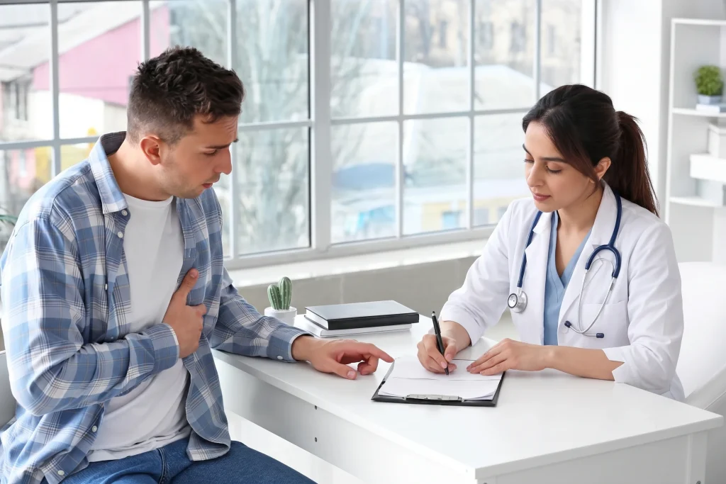 Female doctor taking notes while consulting with a male patient holding his chest at a no insurance medical clinic.
